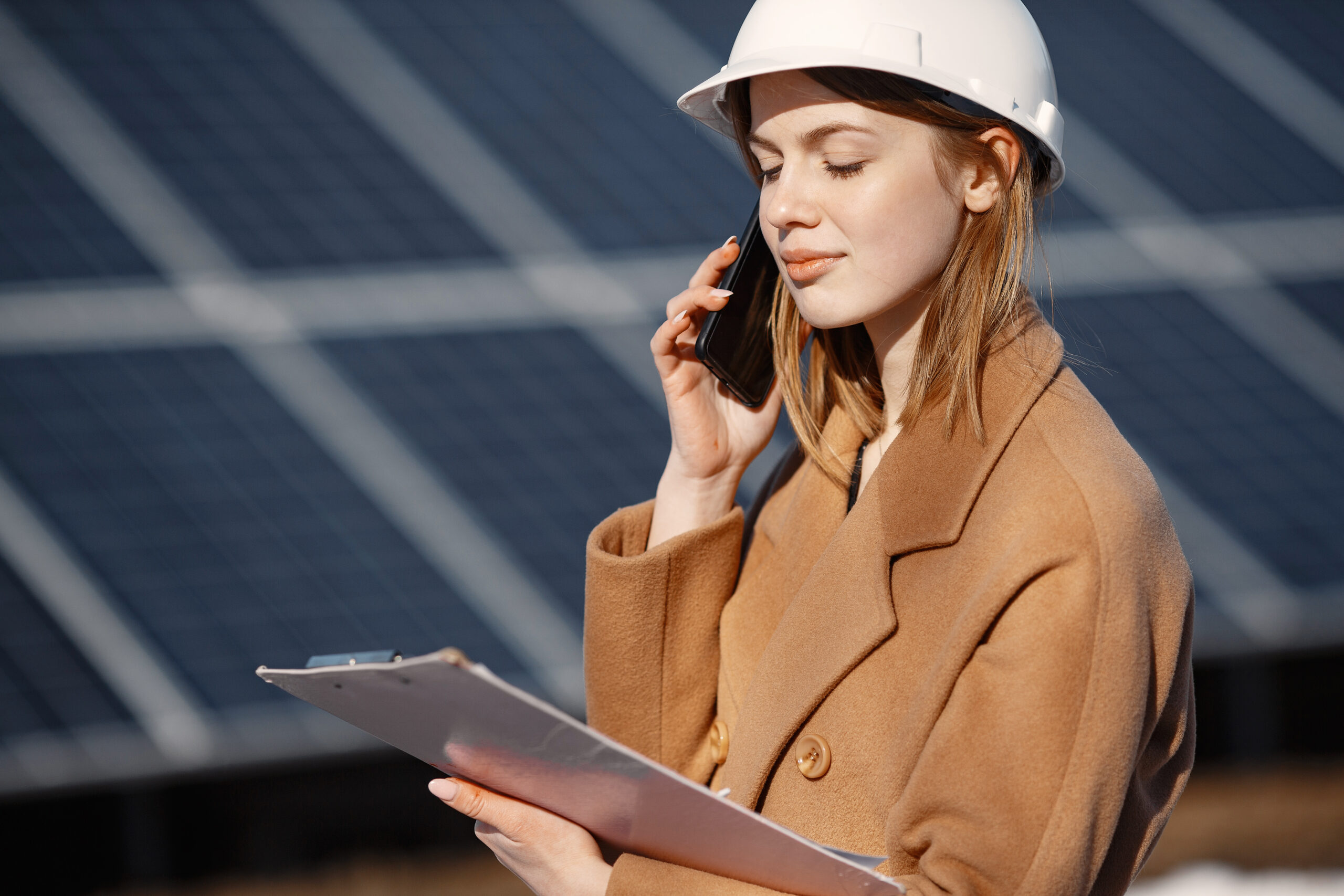 Solar energy station. Young female engineer work at plant. She is talking by phone anddoing business. Woman in helmet with papers.
